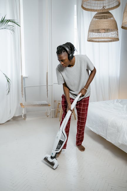 Close-up of a red upright vacuum cleaner being used on a polished wooden floor in a modern room, with visible white drawers and furniture in the background illuminated by natural light. The vacuum cle